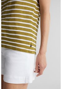 Persona vistiendo pantalones cortos blancos y una camiseta a rayas verdes y blancas, con la mano derecha relajada a un lado contra un fondo liso.
