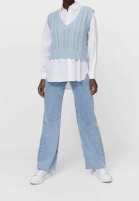 Osoba ubrana w jasnoniebieski sweter w warkocze na białej koszuli z guzikami, jasnoniebieskie spodnie typu wide-leg i białe trampki, stoi z nogami w rozkroku.