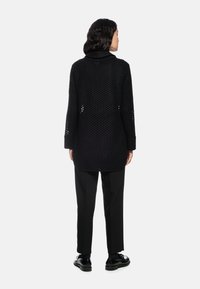 Schwarze Strickjacke mit langem Schnitt und perforiertem Design, langen Ärmeln und hohem Kragen, kombiniert mit schwarzen Hosen und Schuhen.