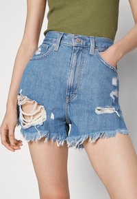 Pantalones cortos de mezclilla en azul claro con detalles desgastados, dobladillo deshilachado y bolsillos visibles. Presenta desgastes en la parte delantera para un aspecto relajado.