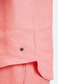 Korallinvärisen pellavapaidan helma, jossa pieni musta pyöreä metallinappi ommeltuna lähelle reunaa.