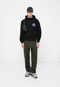 Zwarte hoodie met logo, olijfgroene cargobroek, grijze sneakers en een zwarte pet. Inclusief een kleine zwarte schoudertas.
