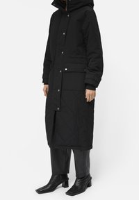 Kvinde iført en lang sort quiltet frakke med trykknapper og sidelommer, sorte cropped bukser og sorte hælankle støvler.