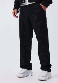 Pantaloni neri con un sottile motivo geometrico, dotati di tasche laterali e un design a gamba dritta, abbinati a scarpe sportive bianche.