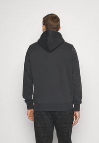 Sweatshirt à capuche gris foncé avec une capuche spacieuse, fabriqué en tissu doux. Présente des poignets et un ourlet côtelés. Porté avec un pantalon noir avec un motif logo.