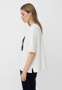 Mujer joven con el cabello lacio de color castaño claro, vistiendo una camiseta blanca suelta con un estampado gráfico y pantalones oscuros, de pie en perfil contra un fondo liso.