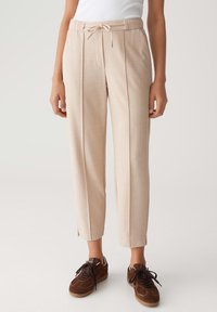 Pantalons tapered beiges taille haute avec plis à l'avant, une ceinture à cordon ajustable, et des baskets à lacets marron portées par une personne debout.