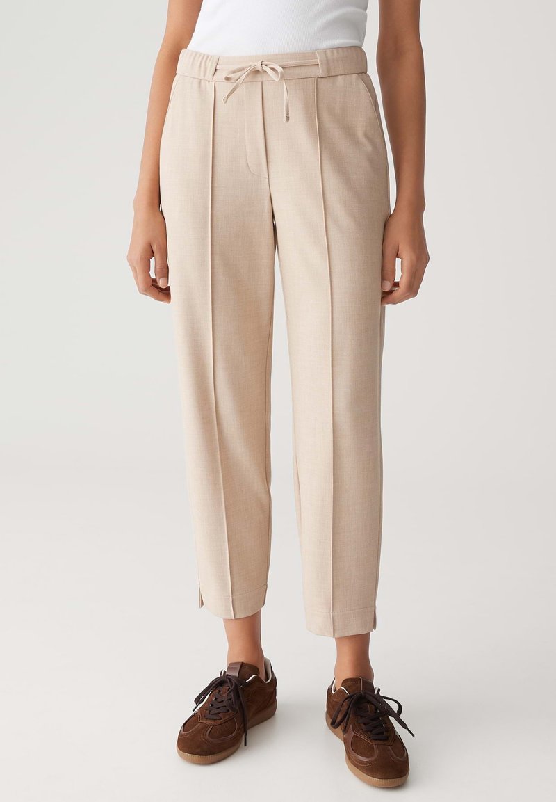 Pantalons tapered beiges taille haute avec plis à l'avant, une ceinture à cordon ajustable, et des baskets à lacets marron portées par une personne debout.