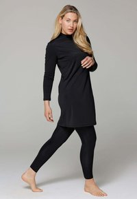 Zwarte langwerpige tuniek met een hoge halslijn, gecombineerd met zwarte leggings. De stof lijkt glad, rekbaar en aansluitend te zijn.