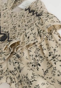 Tissu beige clair à motif floral avec des bords froncés et une texture plissée. Des broderies en fils sombres ajoutent des détails au design.