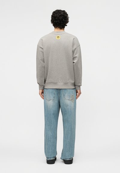 Άτομο με σγουρά μαλλιά στέκεται πλάτη, φορώντας γκρι φούτερ με ένα μικρό κίτρινο γραφικό προσώπου και ανοιχτό μπλε τζιν παντελόνι με φαρδύ πόδι.