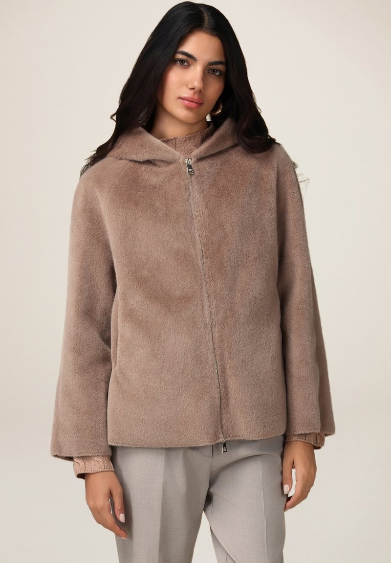 Faux fur zip-jakke i blød beige, med hætte, brede ærmer og en kort kant, parret med lysfarvede bukser.