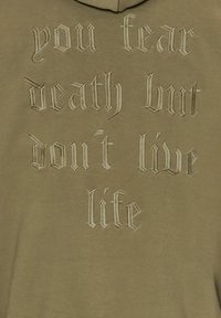 Felpa con cappuccio verde oliva con testo ricamato in beige chiaro, che recita: "temi la morte ma non vivi la vita." Texture liscia.
