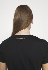 Black cotton t-shirt featuring a round neckline and short sleeves. Notable detail: gold "KARL LAGERFELD" logo on the back.
