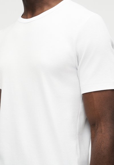 Κοντινό πλάνο ενός ατόμου που φορά ένα απλό λευκό T-shirt με κοντά μανίκια και στρογγυλή λαιμόκοψη, δείχνοντας τον άνω κορμό και το χέρι.