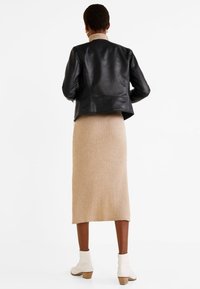 Giacca nera di pelle con silhouette aderente, gonna midi a costine beige e stivaletti bianchi alla caviglia su un modello visto da dietro su uno sfondo neutro.