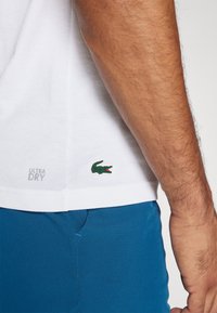 Camiseta blanca de algodón con un logo de cocodrilo verde y el texto "ULTRA DRY" en el dobladillo, combinada con pantalones de tela azul.
