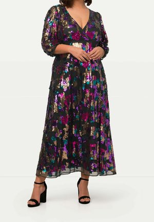 Femme portant une longue robe noire avec des sequins violets, dorés et bleu sarcelle, des manches transparentes et des sandales noires à talons avec des brides à la cheville.