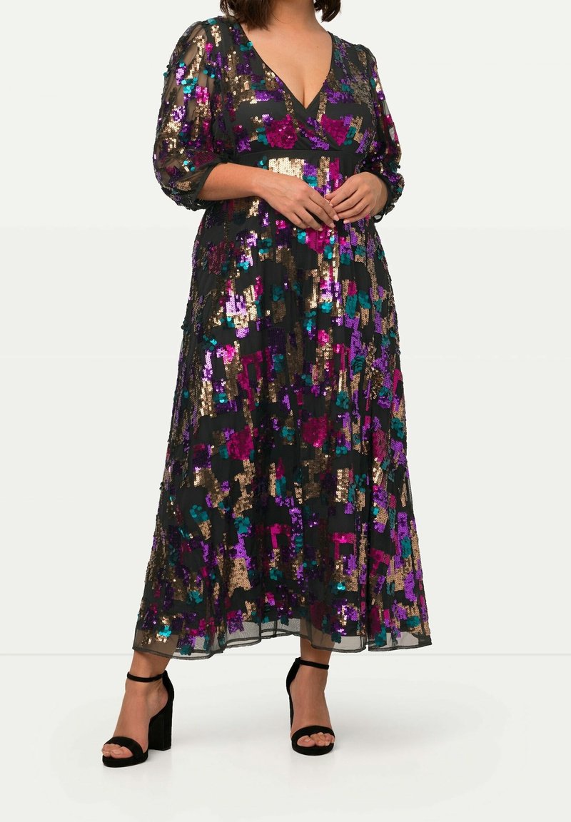 Femme portant une longue robe noire avec des sequins violets, dorés et bleu sarcelle, des manches transparentes et des sandales noires à talons avec des brides à la cheville.