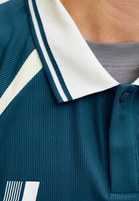 Gros plan sur un polo bleu côtelé avec un col blanc orné de deux rayures bleu marine, porté par-dessus un sous-vêtement gris.