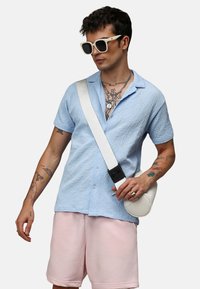 Camisa de manga corta texturizada azul claro, pantalones cortos rosas, bolso cruzado blanco y gafas de sol negras de gran tamaño. Presenta tatuajes visibles.