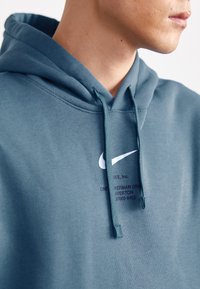 Hoodie i tealfärgat tyg med en vit Nike-logotyp och tryckt text i mörklila. Dragsnörena är matchande teal och har en mjuk textur.