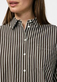 Camicia a righe marroni e bianche con colletto, dotata di taschino frontale e texture liscia. Cinque bottoni visibili lungo la pattina.