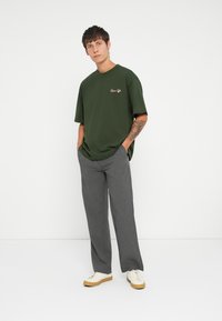 Camiseta oversized verde olivo con un pequeño logo bordado, combinada con pantalones anchos de rayas grises y zapatillas blancas con suelas de goma.