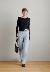 Μαύρη μπλούζα με μακριά μανίκια, ανοιχτό μπλε παντελόνι υψηλής μέσης, μαύρη ζώνη, μαύρη τσάντα χειρός, μαύρες γόβες με τακούνι, ξύλινο πάτωμα, ουδέτερο φόντο.