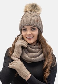 Chapeau beige tricoté avec un pompon en fausse fourrure, gants assortis et une écharpe texturée épaisse. Matériaux chauds et accents côtelés.