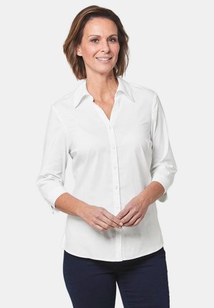 Blusa bianca con colletto e maniche a tre quarti. Tessuto liscio, design aderente e leggero lucido. Indossata con pantaloni blu scuro.