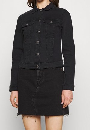 Kobieta ubrana w zapinany na guziki czarny dżinsowy kurtkę z kieszeniami na piersi oraz czarną dżinsową spódnicę z wysokim stanem i postrzępionym dołem.