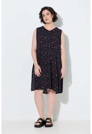 Frau mit kurzen lockigen Haaren, die ein ärmelloses schwarzes Kleid mit rotem Blumenmuster und schwarze Sandalen mit goldenen Schnallen trägt, steht vor einer weißen Wand.