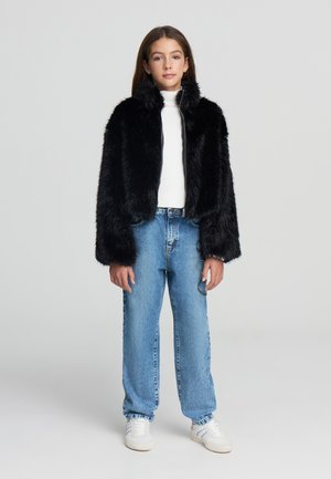 Giovane che indossa una giacca nera in pelliccia sintetica, un dolcevita bianco, jeans blu con dettagli in catena e sneakers bianche, in piedi di fronte a uno sfondo semplice.