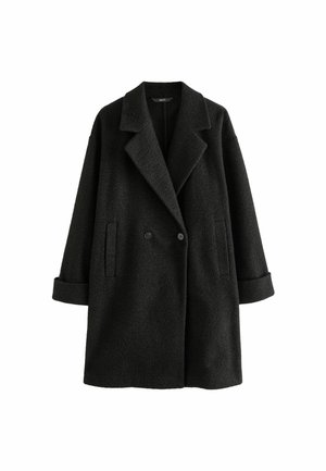 Personne portant un manteau en laine noir ceinturé, un haut blanc, un pantalon imprimé léopard et des ballerines noires, debout contre un mur beige texturé.