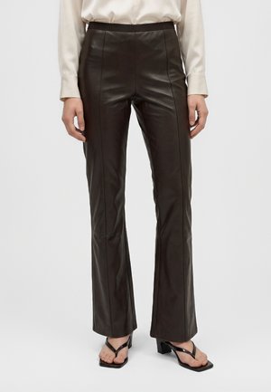Modèle portant un pantalon évasé marron foncé effet cuir, un chemisier crème et des sandales noires sur un fond de studio uni.