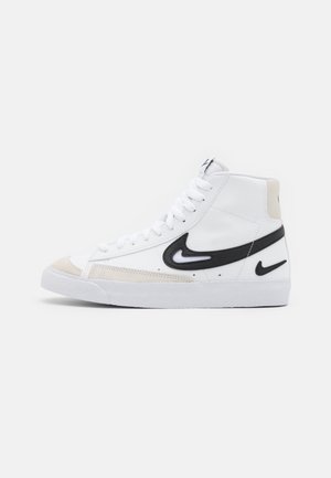 Basket montante blanche avec logos Nike swoosh noirs à double couche, accents beige aux orteils et talons, et lacets blancs sur fond uni.