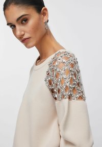 Pulover bej cu design texturat, având un mânecă decorată cu model de diamante cu paiete argintii și o siluetă strâmtă.