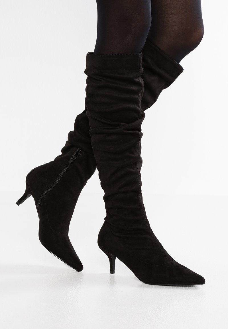 Botas de ante negras hasta la rodilla con un diseño holgado, punta afilada y un tacón delgado. Cuentan con una cremallera lateral para facilitar su uso.