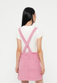Rosa Cord-Röcken Overall-Kleid mit über Kreuz verlaufenden Trägern, zwei Hintertaschen und gesticktem Logo am Bund. Weiße T-Shirt darunter.