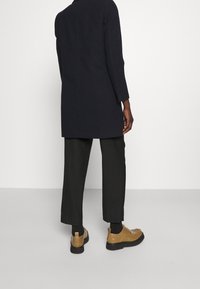 Svart lång kappa med rak skärning, matchad med mörka byxor och bruna snörskor. Skorna har en slät yta och svarta sulor.