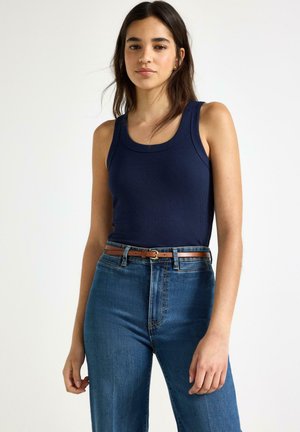 Kvinde med langt mørkt hår iført en navyblå ærmeløs ribstrikket top, højttaljede blå jeans og et brunt bælte, stående foran en ensfarvet baggrund.