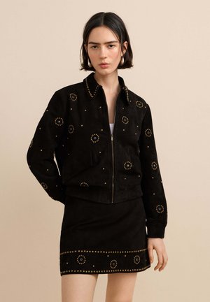 Vrouw met kort donker haar draagt een zwarte jas met ritssluiting en een bijpassende rok, versierd met ronde gouden studs.