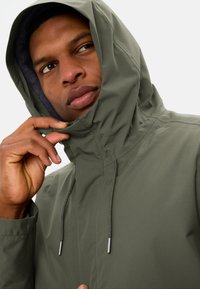 Groene waterdichte jas met een capuchon, voorzien van verstelbare trekkoorden en een zachte voering, gedeeltelijk dichtgeritst bij de kraag.