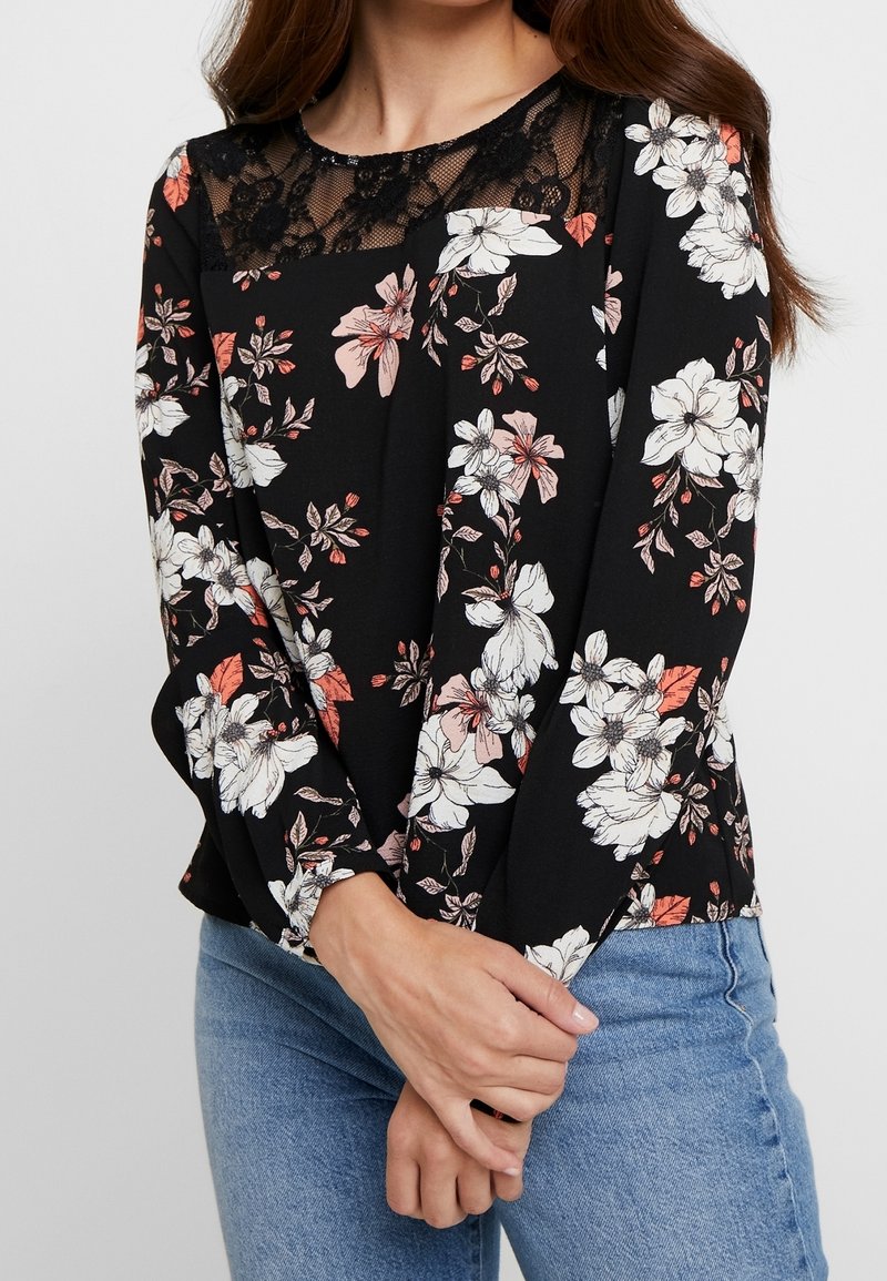 Blouse noire à fleurs avec manches longues, détail en dentelle au col, et un motif de fleurs roses et blanches, portée avec un jean bleu.