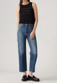 Kvinna i svart ärmlös virkat topp, högmidjade blå raka jeans och svarta chunky loafers, stående mot en enkel bakgrund.