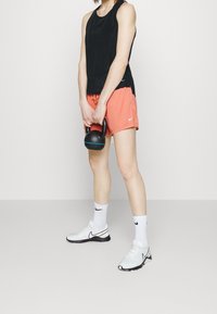 Osoba w czarnym topie na ramiączkach, pomarańczowych szortach, białych skarpetkach i butach trzymająca oburącz ketlebell przed nogami.