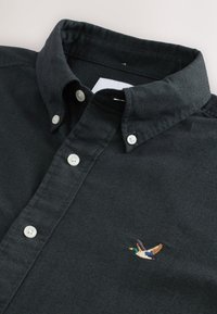 Čierna košeľa na zapínanie s golierom, bielymi gombíkmi a vyšitým motívom kačky na ľavej strane hrudníka. Bavlnená textúra.