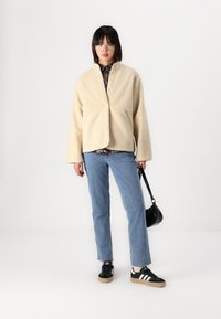 Beige fleecejakke med knaplukning, oversized pasform, parret med lyseblå lige ben jeans og sorte sneakers med hvide striber.