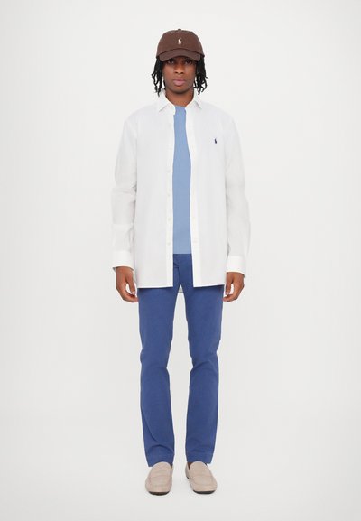 Camicia bianca con bottoni sopra una t-shirt blu, pantaloni blu e un cappello marrone con un logo. Mocassini neutri completano l'outfit.